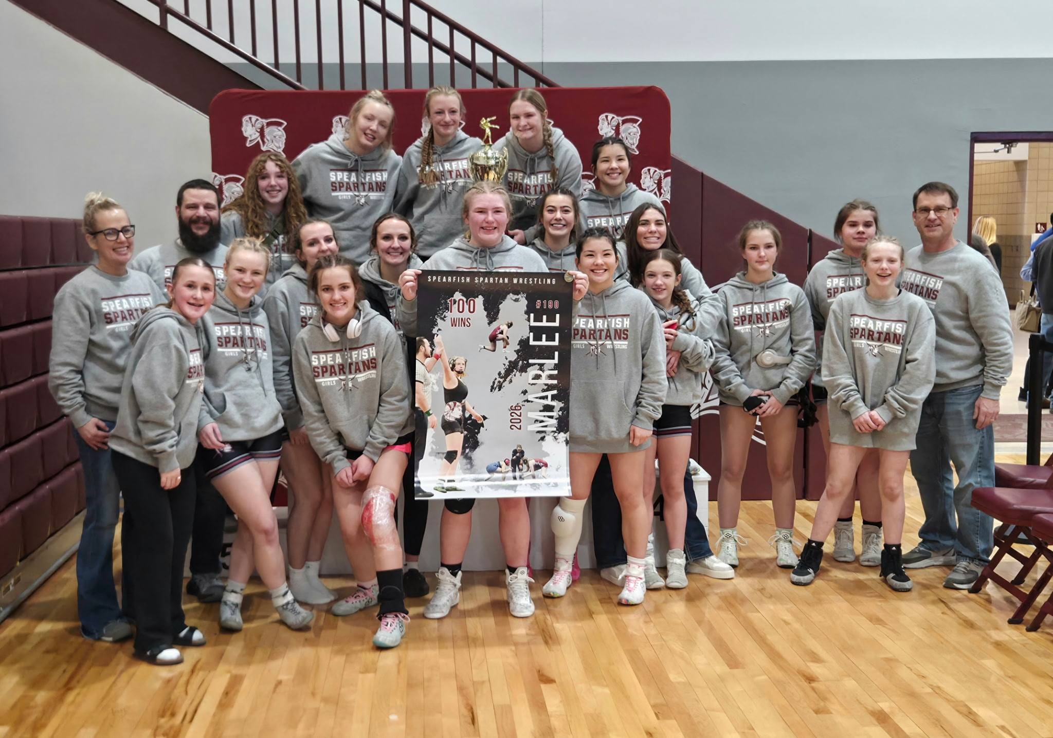 Spearfish Spartans girls wrestling team and coaches pose in the gym with a conference trophy and a “100 Wins” poster, celebrating their fifth consecutive Black Hills Conference team championship and Marlee Heltzel’s 100th career varsity win.