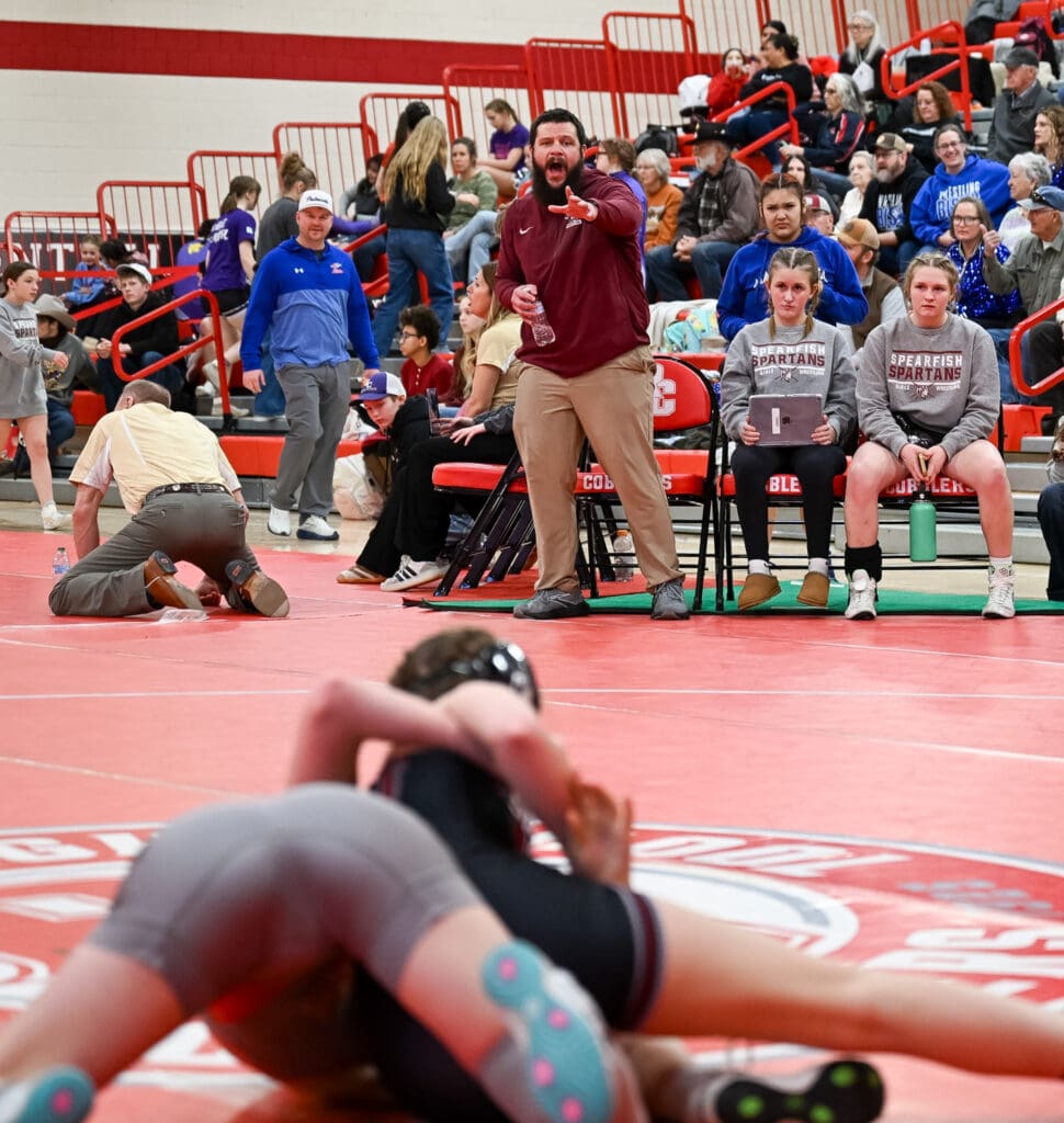 Spearfish High School girls wrestling head coach Cody Powers giving instructions to a Spartan wrestler during a match, with a scoreboard and crowd in the background.